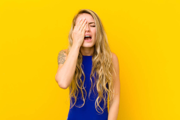 young blonde woman looking sleepy, bored and yawning, with a headache and one hand covering half the face against yellow wall - Foto, Bild