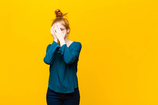 young red head woman covering face with hands, peeking between fingers with surprised expression and looking to the side against orange wall - Foto, imagen