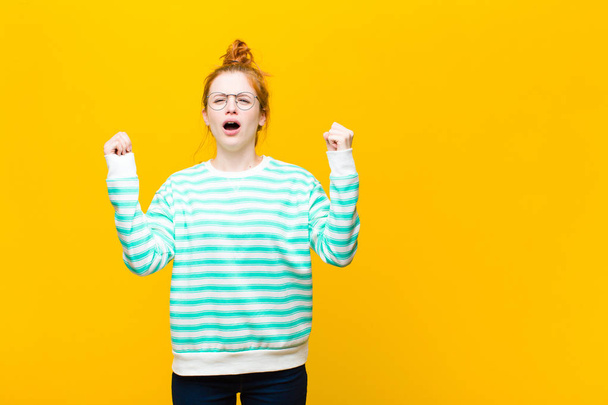 young red head woman shouting aggressively with an angry expression or with fists clenched celebrating success against orange wall - Foto, Imagen
