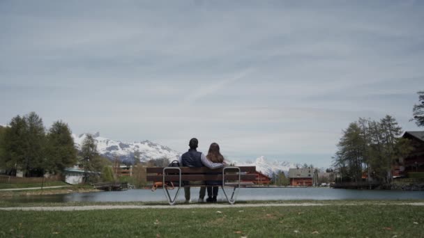Rear view of romantic young couple sitting on bench at lakeside with scenic mountain view. General plan - Кадры, видео