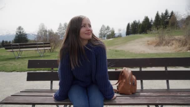Smiling young woman sits on a wooden bench with amazing view over lake and mountains and relaxed on sunny spring day. Woman enjoying a picturesque place. - Video