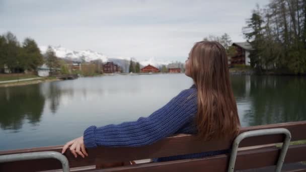 Young woman sits on a wooden bench with amazing view over lake and mountains and relaxed on sunny spring day. Woman enjoying a picturesque place. Rear view - Felvétel, videó