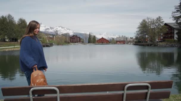 Šťastná žena přichází k dřevěné lavičce s úžasným výhledem na jezero a hory a sedí. - Záběry, video