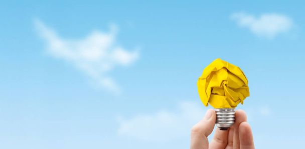 Business Idea Concept : Hand holding light bulb made from yellow crumpled paper ball with blue sky in background. - Photo, Image