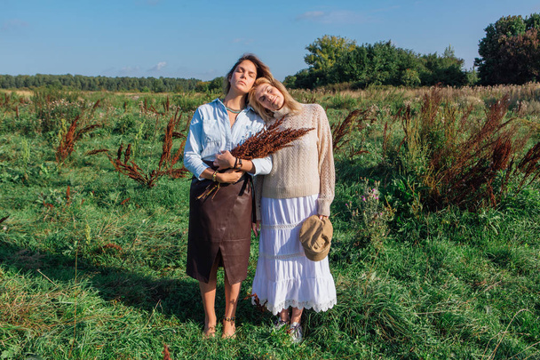 Porträt zweier schöner Frauen auf einem Feld im Spätsommer. schöne Mädchen, die nebeneinander stehen und einen Strauß aus trockenen braunen Pflanzen in der Hand halten. - Foto, Bild