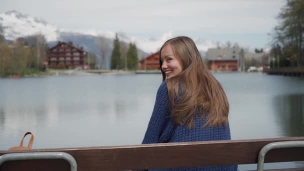 Beautiful long-hair woman sitting back on wooden bench at lakeside with scenic mountain view. Then girl looking back and smiling beautifully to the camera - Πλάνα, βίντεο