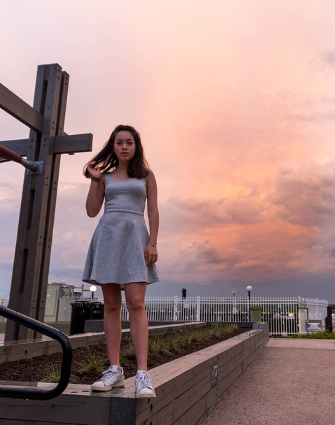Asian teen in grey dress standing on wall with sunset in the background - Fotoğraf, Görsel
