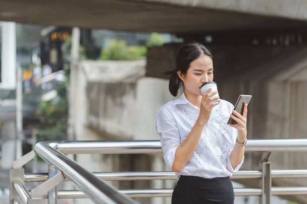 若い女性の白いシャツ立ち飲みコーヒーと仕事のためのスマートフォンを使用して. - 写真・画像