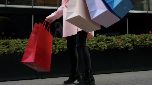 Jonge aantrekkelijke vrouw is winkelen. Dame met papieren zakken in haar handen, dansen op straat tegen de achtergrond van groene bloembed van de gevel van het winkelcentrum. Kerst vakantie, verkoop, Retail industrie concept. - Video