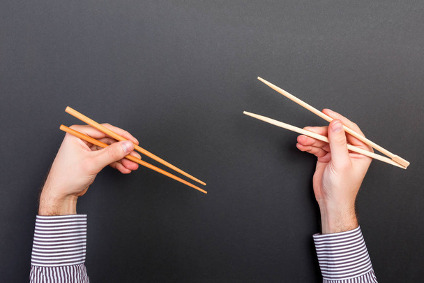 Kreatives Bild von Holzstäbchen in zwei Männerhänden auf schwarzem Hintergrund. Japanisches und chinesisches Essen mit Kopierraum - Foto, Bild