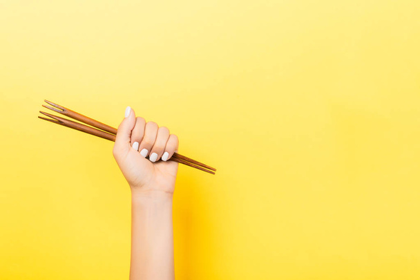 Cropped image of female hand holding chopsticks in fist on yellow background. Asian food concept with copy space - Valokuva, kuva