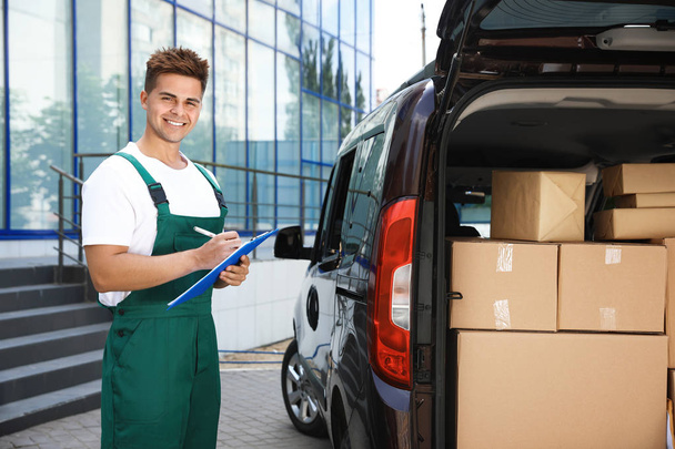 Young courier holding clipboard near delivery van with parcels outdoors - Foto, imagen