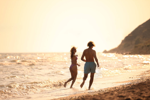 Young woman in bikini and her boyfriend having fun on beach at sunset. Lovely couple - Zdjęcie, obraz