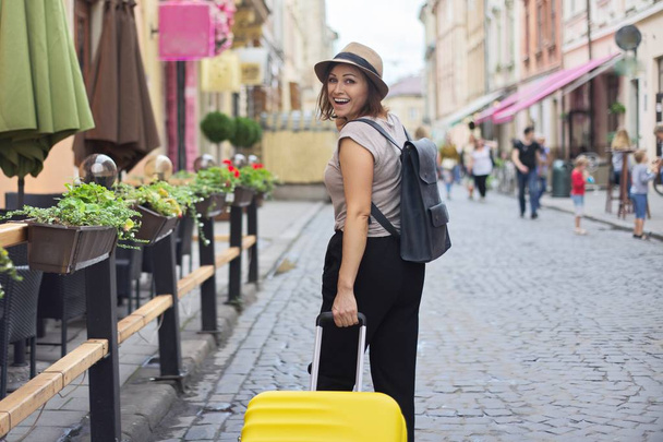 Mature smiling woman traveling in tourist city with suitcase - Fotó, kép
