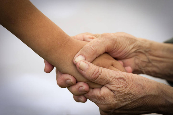 Τα χέρια ενός ηλικιωμένου ανθρώπου κρατούν τα χέρια ενός παιδιού. Η έννοια της φροντίδας και της βοήθειας για τους ηλικιωμένους, η σύνδεση της παλαιότερης και νεότερης γενιάς των ανθρώπων. Εικόνα. - Φωτογραφία, εικόνα