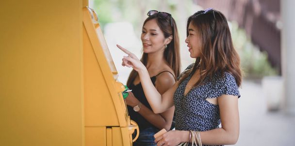 Dwie piękne azjatyckie kobiety wycofując pieniądze z karty bankowej za pomocą bankomatu w centrum handlowym  - Zdjęcie, obraz