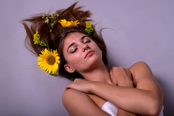 Portrait of a beautiful girl lying with sunflower flowers in her hair with closed eyes in a studio on a gray background. - Photo, image