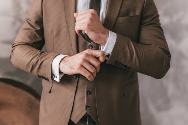 Elegant young fashion man looking at his cufflinks while fixing them. Photo of male hands. Handsome groom dressed in brown formal suit, white shirt and tie is getting ready for wedding - Fotó, kép