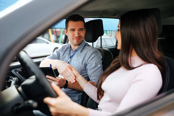Male auto instructor takes exam in young woman. Shaking hands. Sitting in car. Brunette hold hand on steering wheel. - Fotografie, Obrázek