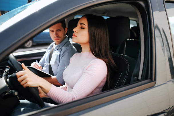 Male auto instructor takes exam in young woman. She hold hands on steering wheel and driving. Young man look at brunette with confusion. Model drives straight. Sit in car. - Фото, зображення