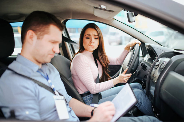 Male auto instructor takes exam in young woman. Worried young woman look at test results. Holding hands on steering wheel. Confused man look down. Daylight. - Foto, immagini