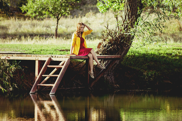 cena romântica de jovem mulher sentada em uma ponte fluvial de madeira com um livro nas proximidades e apreciando a natureza do outono, menina relaxante andando ao ar livre
 - Foto, Imagem