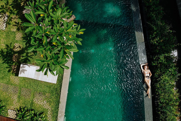 Top view of woman in beige bikini relaxing on edgeof infinity pool around palm trees and jungle. Luxury holiday.Girl resting on the island of Bali. Copy space. Vacation concept - Fotoğraf, Görsel