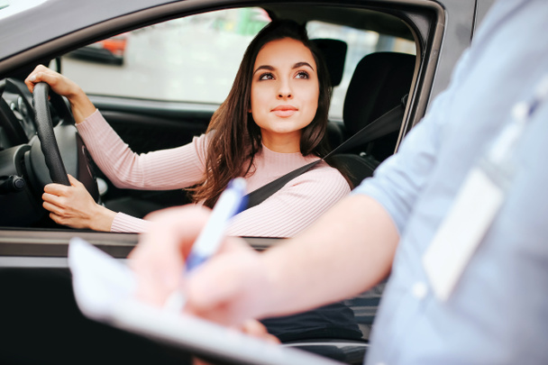 Male auto instructor takes exam in young woman. Guy writes test result in paper. Interesting woman look at guy outside from car. Hold hands on steering wheel and seat belt across body. - 写真・画像