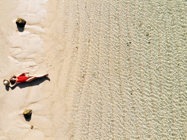 Aerial drone view of young woman in red bikini sunbathe on beach with white sand, turquoise water of the Indian Ocean. Bali Island, Indonesia. Tropical background and travel concept - 写真・画像