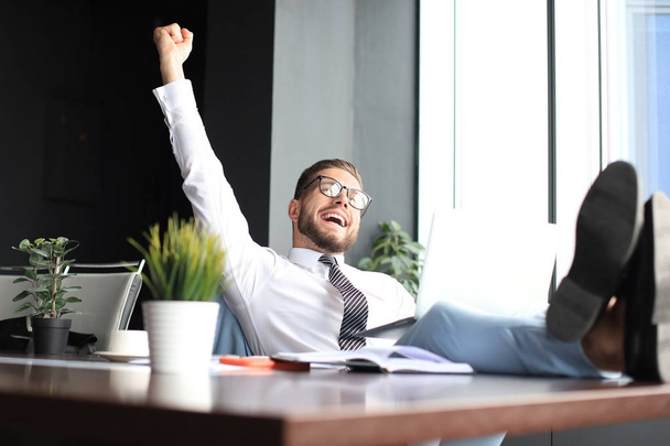 Handsome businessman sitting with legs on table and keeping arm raised and expressing joyful in office - Foto, Bild