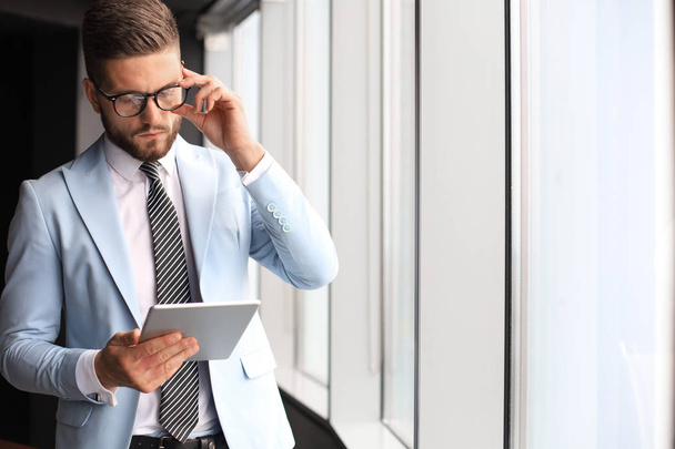 Modern business man in formalwear using digital tablet while standing near window in the office - Фото, изображение