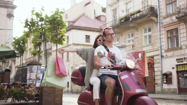 Adorable happy young lady in stylish sunglasses with purchases riding on the scooter with her boyfriend through the urban street - Footage, Video