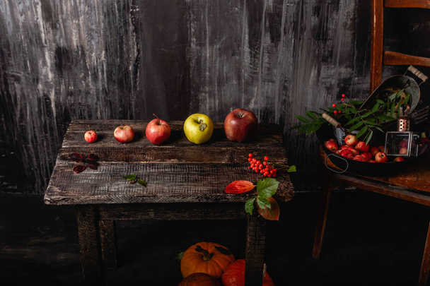 collection of apples on wooden table and chair with pumpkins on floor in dark room - Zdjęcie, obraz