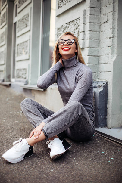 young girl in glasses,wearing gray jeans and gray sweater. sitting on the pavement and smile. Pretty laughing girl has a good time in autumn weekend - Foto, afbeelding