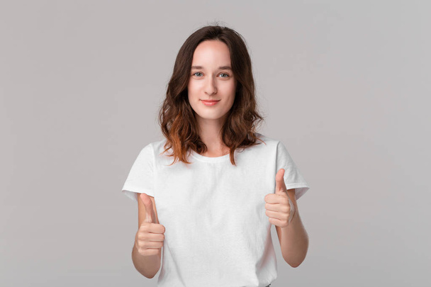 Pretty brunette woman in white t-shirt holding thumbs up expressing positive evaluation standing isolated over grey background. I like it! - Photo, Image