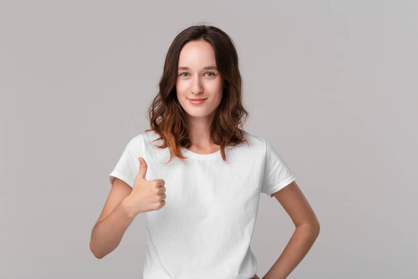 Pretty brunette woman in white t-shirt holding thumbs up expressing positive evaluation standing isolated over grey background. I like it! - Photo, Image