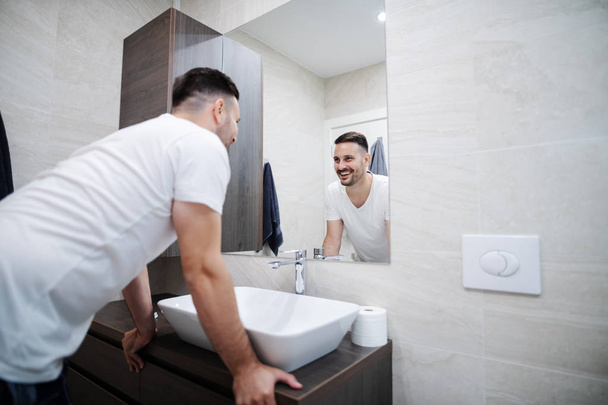 Knappe Kaukasische glimlachend in pyjama staande in moderne badkamer en op zoek naar zichzelf in spiegel. Ochtend tijd. - Foto, afbeelding