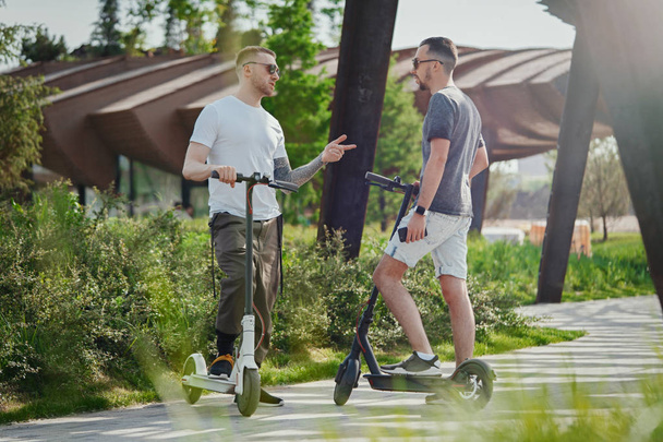 Two handsome men talking next to electric kick scooters at park landscape - Фото, изображение