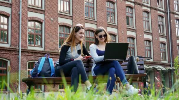 Moderne Studentinnen arbeiten gemeinsam an einem Laptop an einem Studentenprojekt, während sie vor dem Studium auf einer Bank im Campus-Park sitzen. Hochschulbildung - Filmmaterial, Video