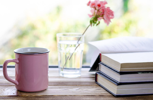 A rózsaszín csésze tea áll egy fa ablakpárkányon a könyvek mellett, és egy pohár, amelyben áll egy rózsaszín muskátli virág - Fotó, kép