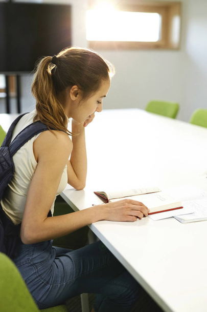 Jeune étudiante enthousiaste d'une école d'informatique assise à table dans une salle de classe, lisant des notes et des informations pour un cahier d'exercices - Photo, image