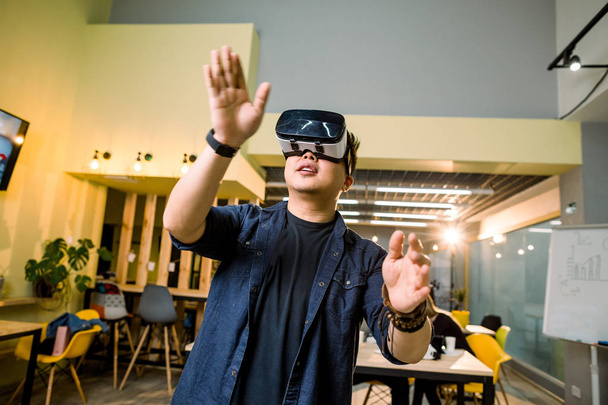 young asian man in casual clothes wearing head-mounted virtual reality goggles. Man with vr glasses posing in modern studio coworking - Foto, imagen