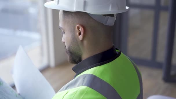 Close-up of bearded worker in helmet and green jacket reading documents. Professional builder with building drawings in hands. Job, profession - Imágenes, Vídeo