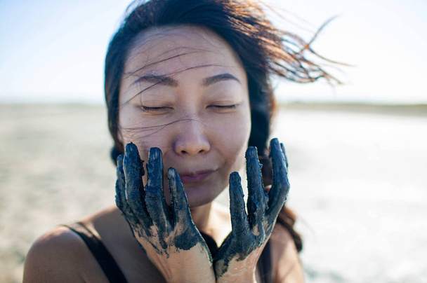 Fiatal gyönyörű ázsiai nő alkalmazó fekete kozmetikai maszk az arca a terápiás fekete sár. Gyógykezelés és arcszépészeti koncepció. Női Arcápolás és tökéletes bőr koncepciók. - Fotó, kép
