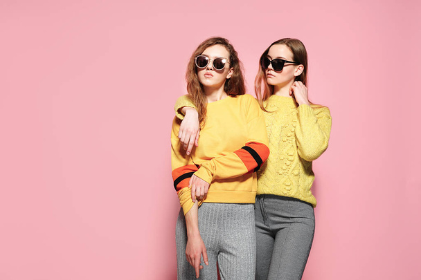 Two beautiful gorgeous girls standing in stylish yellow sweaters, gray pants, sunglasses on pink background. - Fotó, kép
