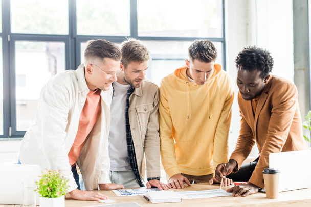 vier junge multikulturelle Geschäftsleute analysieren Aufsätze mit Diagrammen und Diagrammen, während sie gemeinsam im Büro an einem Startup-Projekt arbeiten - Foto, Bild