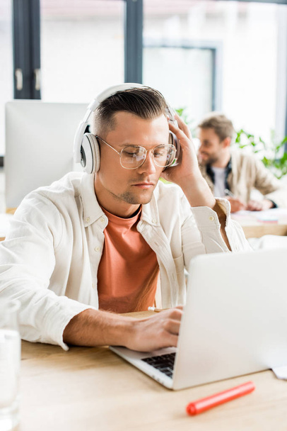 young pensive businessman using laptop while sitting in headphones - Φωτογραφία, εικόνα