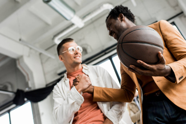 low angle view of african american businessman holding volleyball and shaking hands with colleague in office - Fotografie, Obrázek