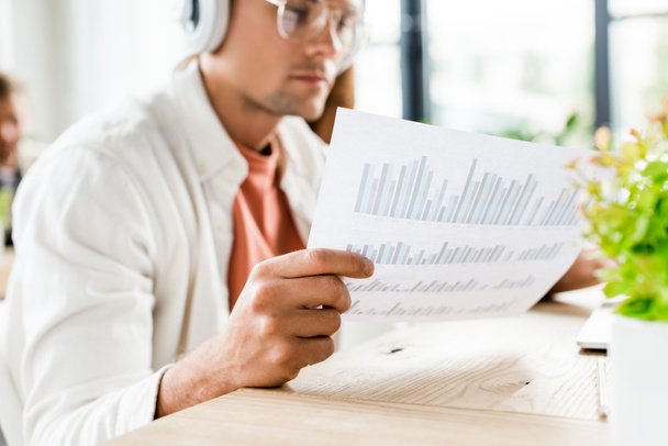 selective focus of young businessman sitting in headphones and analyzing paper with graphs and charts - Fotó, kép