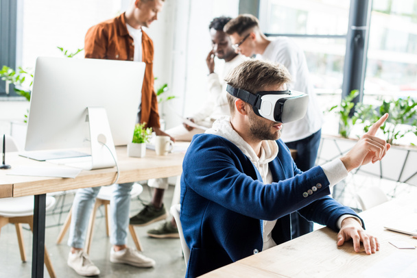 young businessman using vr headset and touching something with finger while multicultural colleagues working in office - Foto, immagini